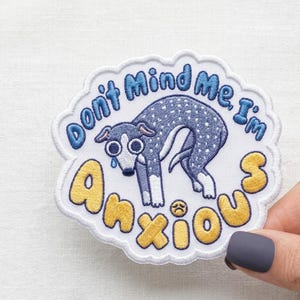 May include: Embroidered patch with a white border and the text "Don't Mind Me, I'm Anxious" in blue and yellow. The patch features a cartoon dog with a sad expression, tears, and a worried emoji.