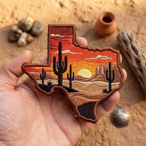 May include: A Texas-shaped embroidered patch featuring a desert landscape at sunset. The design includes black silhouettes of saguaro cacti against a vibrant orange and red sky, a yellow sun, tan desert terrain, and a dark brown border.