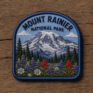May include: Embroidered patch featuring Mount Rainier National Park. The design includes a snow-capped mountain, evergreen trees, wildflowers, and the text "Mount Rainier National Park" in a blue arched border.