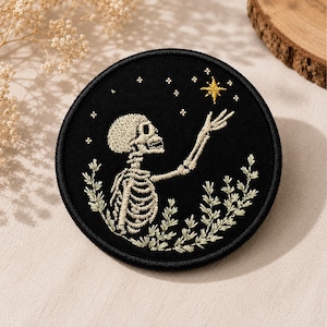 May include: A circular embroidered patch featuring a black background with a white skeleton reaching toward a gold star. The skeleton is surrounded by white botanical sprigs and small white stars scattered across the upper portion of the patch.