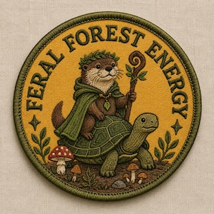 May include: Embroidered patch with a whimsical design. A brown otter wearing a green cape and laurel wreath rides a green turtle, holding a staff. The patch reads "FERAL FOREST ENERGY" in yellow, with forest elements.