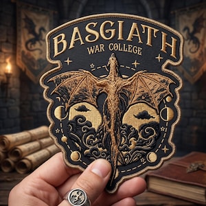 May include: Embroidered patch featuring a golden dragon with outstretched wings, the text "BASGIATH WAR COLLEGE" in gold lettering, and celestial elements. The patch is black with gold and cream-colored details. The patch is held in a hand.
