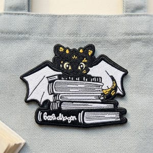 May include: Embroidered patch of a black dragon with white wings on a stack of books. The dragon has gold detailing. The patch includes the text "books dragon" in white. The patch is on a grey fabric background.