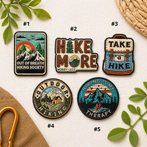 May include: Five embroidered hiking-themed patches with various designs. The patches display phrases such as "Out of Breath Hiking Society," "Hike More," and "Take a Hike." The colour scheme includes greens, blues, oranges, and browns, with images of mountains, trees, and a backpack.