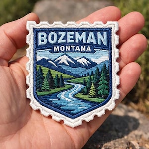 May include: Embroidered patch featuring a scenic landscape with the text "BOZEMAN MONTANA". The design includes mountains, a river, and evergreen trees, all within a blue and white border. The patch is held in a hand.