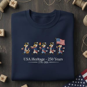 May include: Navy blue sweatshirt with embroidered eagles in colonial attire, marching with a drum, flute, torch, and American flag. The text "USA Heritage - 250 Years 1776-2026" is printed below the eagles. A patriotic design.