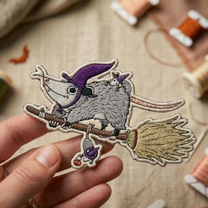 May include: Embroidered patch featuring a gray opossum wearing a purple witch hat, riding a broom. The opossum has a long pink tail and is accompanied by two smaller opossums. The patch is detailed with various colors and textures.