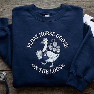 May include: Navy blue sweatshirt with the text "FLOAT NURSE GOOSE ON THE LOOSE" in white. The design features a cartoon goose wearing a nurse's hat and carrying a clipboard. The goose has a badge that says ER, ICU, Med-Surg, and Tele-Metro.