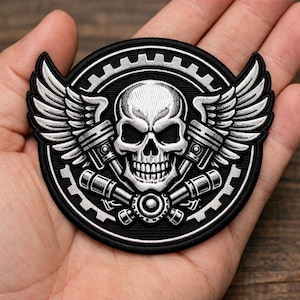 May include: A circular embroidered patch featuring a central white skull with black eye sockets and nose cavity. Behind the skull are two crossed silver pistons and a gear border. Two silver wings extend from the sides of the gear.