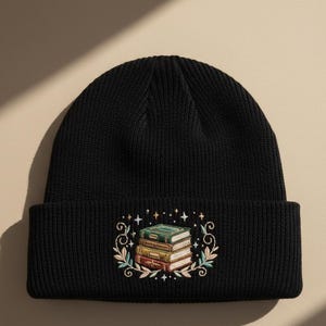 May include: Black knit beanie featuring an embroidered design of a stack of books. The embroidery includes green, gold, and red book spines, surrounded by a floral and star design. The beanie is a classic, comfortable style.