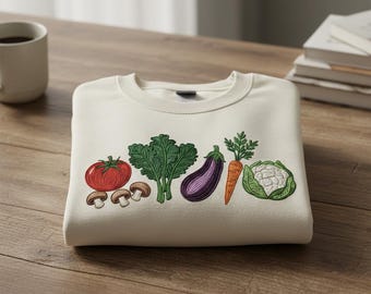 May include: Cream-coloured sweatshirt featuring embroidered vegetables: a red tomato, mushrooms, green kale, a purple aubergine, an orange carrot, and a white cauliflower. The crew neck sweatshirt is folded on a wooden surface.