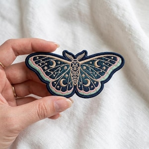 May include: Embroidered moth patch in navy blue, teal, and pink. The patch features a detailed design of a moth with crescent moons and stars. The patch is held in a hand against a white background.