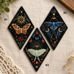 May include: Three diamond-shaped embroidered patches with moth and celestial designs. Each patch has a black background. One features a sun and a moth, another a blue moth, and the third a green moth. Floral and star details are included on each patch.
