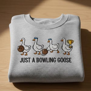 Puede incluir: Sudadera gris claro con la frase "JUST A BOWLING GOOSE" impresa en negro. El dise&ntilde;o presenta gansos de dibujos animados con bolas de bolos y un trofeo.