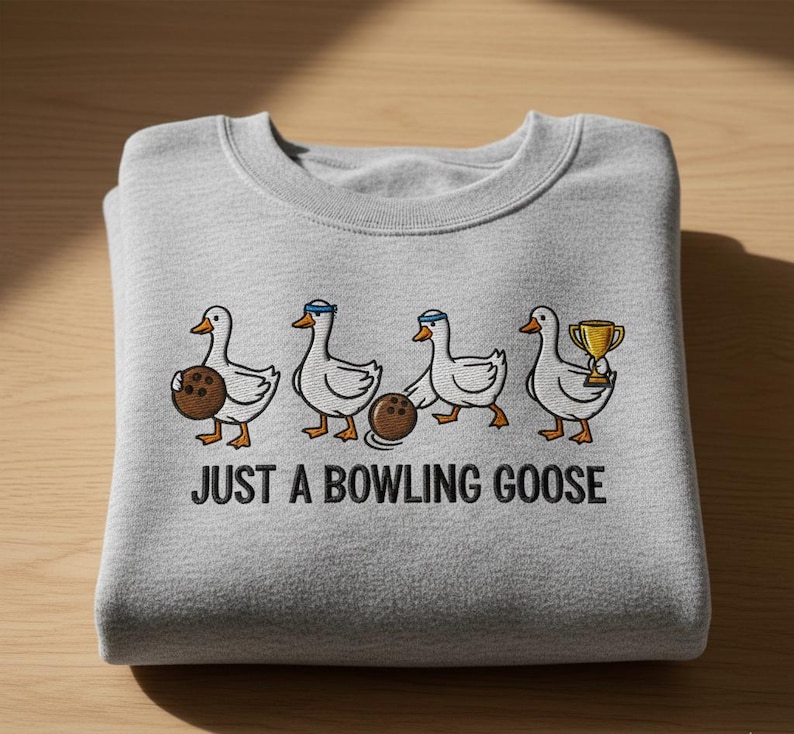 Puede incluir: Sudadera gris claro con la frase "JUST A BOWLING GOOSE" impresa en negro. El dise&ntilde;o presenta gansos de dibujos animados con bolas de bolos y un trofeo.