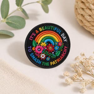May include: Circular black patch with the text "IT'S A BEAUTIFUL DAY TO SMASH THE PATRIARCHY". The patch features a rainbow, colourful flowers, and green leaves.