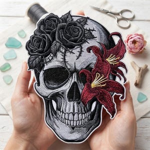 May include: An embroidered patch featuring a detailed gray and white skull adorned with black roses and thorny vines at the top, and two deep red lilies with yellow centers on the right side. The design is outlined in white on a dark background.