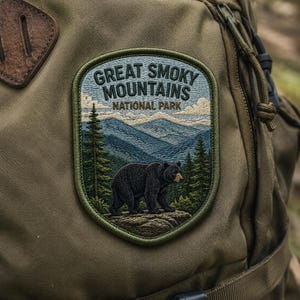 May include: Embroidered patch featuring the text "Great Smoky Mountains National Park." The patch depicts a black bear standing on a rock, with a backdrop of mountains and trees. The patch is attached to a green backpack.