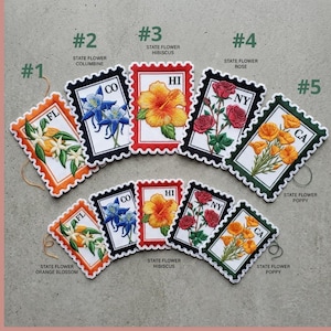 May include: Embroidered patches shaped like postage stamps, each depicting a state flower. The patches showcase the orange blossom, columbine, hibiscus, rose, and poppy. Each patch has a unique colour scheme and state abbreviation.