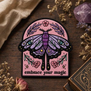 May include: Embroidered patch featuring a purple moth with intricate details, set against a pink background. The design includes floral and celestial elements, with the words "embrace your magic" at the bottom. The patch is framed by a black border.