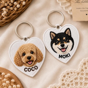 May include: Two heart-shaped keychains with embroidered dog faces. One features a brown poodle with the name "COCO", and the other a black and tan Shiba Inu with "MOKI". Both have silver key rings and white borders.