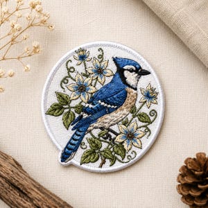 May include: Embroidered patch featuring a blue jay perched among blue and cream-colored flowers and green leaves. The patch is circular with a white border. The bird has blue, black, and tan feathers. Ideal for jackets, bags, or other fabric items.
