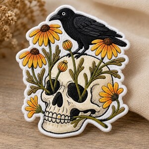 May include: An embroidered patch featuring a cream-colored human skull with a black crow perched on top. Yellow and orange daisy-like flowers with green stems grow from the eye sockets and mouth of the skull, surrounded by a white border.