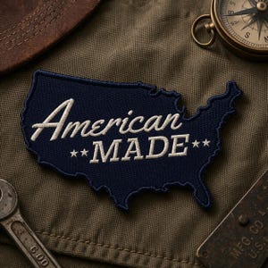 May include: A navy blue patch in the shape of the United States with the words "American MADE" embroidered in white. The patch is on a green fabric background, with a leather hat, compass, and wrench visible.