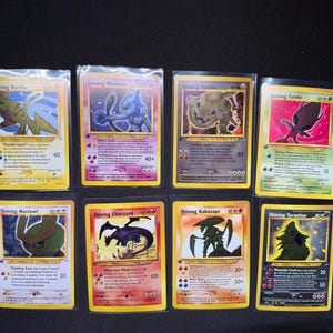 Puede incluir: Ocho cartas coleccionables de Pokémon, cada una en fundas protectoras, se muestran sobre un fondo negro. Las cartas presentan varios personajes de Pokémon, incluyendo Shining Raichu, Mewtwo, Charizard, y otros, con colores vibrantes y diseños únicos.