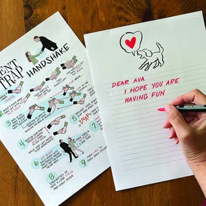 May include: Two sheets of white paper on a wooden surface. One sheet has instructions for "The Parent Trap Handshake." The other has a dog illustration with a heart and the text "Dear Ava, I hope you are having fun." A hand is writing on the second sheet with a red marker.