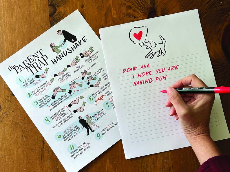 May include: Two sheets of white paper on a wooden surface. One sheet has instructions for "The Parent Trap Handshake." The other has a dog illustration with a heart and the text "Dear Ava, I hope you are having fun." A hand is writing on the second sheet with a red marker.