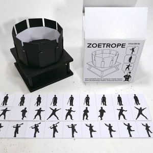 Puede incluir: Un zoótropo en blanco y negro con un cilindro de papel blanco y una base negra. El zoótropo está etiquetado como "ZOETROPE 1914/28/36". Hay 18 imágenes en blanco y negro de una figura de palo en tarjetas blancas.