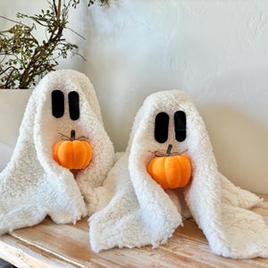 May include: Two white fuzzy ghost decorations with black eyes and a small orange pumpkin in their hands. The ghosts are sitting on a wooden surface.