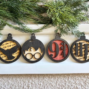 May include: Four wooden Christmas ornaments with black outlines and various designs. One features a silhouette of a bird, another glasses, the third the numbers 9 3/4, and the last the letters HP with a lightning bolt. The ornaments are on a white tray.