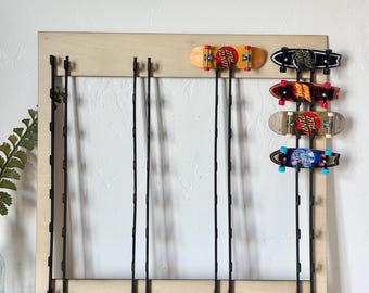 May include: A wooden wall-mounted rack with black metal hooks for displaying fingerboard skateboards. The rack has five hooks and is holding four fingerboards, including a black and white fingerboard with "Santa Cruz" written on it, a red and black fingerboard with "Santa Cruz" written on it, a black and yellow fingerboard with "Santa Cruz" written on it, and a blue and white fingerboard with "Santa Cruz" written on it.