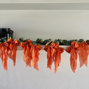 May include: A shelf decoration featuring a garland adorned with orange fabric bows and green foliage. A black jar with a handle is on the left. The bows are evenly spaced, creating a visually appealing and festive display.
