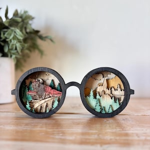 May include: Wooden, black-framed glasses with a layered, laser-cut design. One lens depicts a red train on a bridge, the other a castle and trees. The artwork is set against a light wood background.