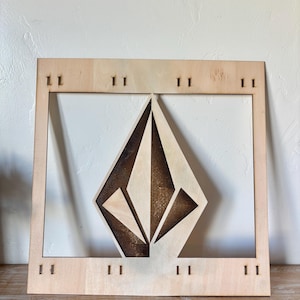 May include: A wooden wall art piece featuring a geometric diamond-shaped design. The artwork is made of light-colored wood with a darker, burnt-wood effect within the diamond. The piece is framed with a rectangular border and has small, evenly spaced cutouts.