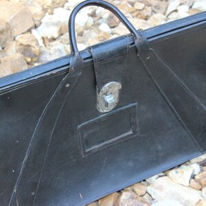 May include: A black briefcase with a textured surface, a single top handle, and a central silver-toned metal latch closure. The rectangular bag features a stitched design with angled panels and a small rectangular identification slot on the front.