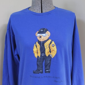 May include: Royal blue long-sleeve shirt featuring a graphic of a bear wearing a yellow and black jacket, black pants, and boots. The bear is also wearing a hat and goggles. The text "POLO BEAR by RALPH LAUREN" is printed below the graphic.