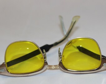 May include: Yellow-tinted sunglasses with a silver frame. The lenses are a bright yellow, and the frame has a classic, rectangular shape. The temples are a light green colour. The sunglasses are designed for eye protection.