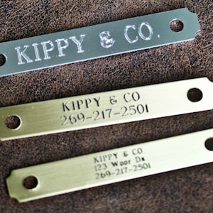 May include: Three metal dog tags with rounded edges. The top tag is silver and says "KIPPY & CO.". The middle tag is gold and says "KIPPY & CO 269-217-2501". The bottom tag is gold and says "KIPPY & CO 123 Woor Da 269-217-2501".