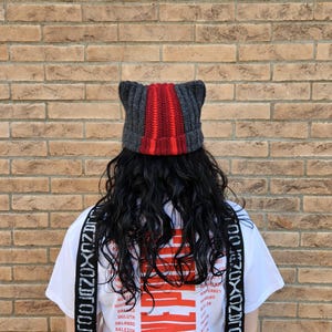 Può includere: Un berretto lavorato all'uncinetto grigio e rosso con orecchie da gatto. Il cappello presenta un design a righe rosse e grigie, con le orecchie da gatto in piedi. La persona indossa una maglietta bianca con un design grafico e bretelle nere.