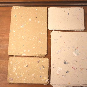 May include: Four stacks of handmade paper with flecks of color. Two stacks are a light tan color, and two are off-white. The paper has a rough, textured appearance, suggesting a natural, eco-friendly product.