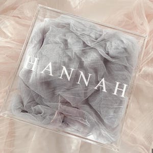 Könnte beinhalten: Eine transparente Acrylbox mit dem Namen "HANNAH" in weißen Buchstaben. In der Box befindet sich ein grauer, durchsichtiger Stoff. Die Box steht auf einem rosa-weißen Stoffhintergrund.