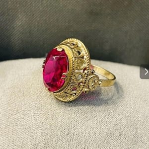 May include: A gold ring featuring a large, oval-cut, vibrant red gemstone set in a decorative, filigree-style metal mounting. The band is plain and circular, while the setting displays intricate, openwork metal patterns surrounding the stone.