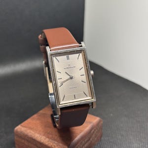 Könnte beinhalten: Eine rechteckige silberne Uhr mit einem braunen Lederarmband. Das Zifferblatt der Uhr ist weiß mit schwarzen römischen Ziffern als Stundenmarkierungen und einem schwarzen Sekundenzeiger. Die Uhr steht auf einem Holzständer.