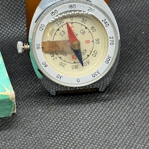 May include: A vintage compass with a silver-colored metal casing and a cream-colored dial. The dial features black and red markings, including degree indicators and directional notations. The compass has a red and black needle and a small winding knob.