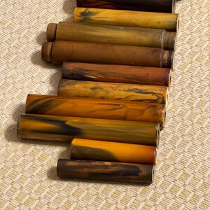 Puede incluir: Una colección de objetos cilíndricos con una variedad de colores, incluyendo tonos de marrón, amarillo y negro. Los objetos tienen una apariencia veteada o arremolinada, y están dispuestos sobre una superficie texturizada.