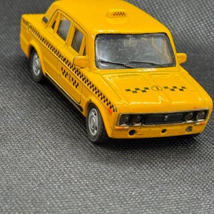 Puede incluir: Un taxi de juguete amarillo con detalles a cuadros negros y la palabra "WOW" en el lateral. El coche en miniatura tiene una luz de techo y una parrilla delantera. El taxi es un modelo vintage, probablemente un objeto de colección.